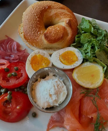 Brunch plate with toasted bagel, whipped cream cheese, smoked salmon (lox), soft‑boiled eggs, tomato slices with capers, pickled red onion, arugula and lemon wedge.