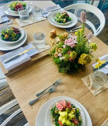 Rustic wooden table set for four with white plates of fresh salads topped with sliced pink seared tuna, glasses of water, folded white napkins and textured silver cutlery, centered around a cheerful pink-and-yellow spring floral centerpiece.