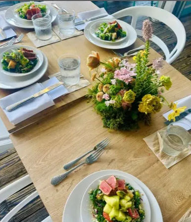Rustic wooden table set for four with white plates of fresh salads topped with sliced pink seared tuna, glasses of water, folded white napkins and textured silver cutlery, centered around a cheerful pink-and-yellow spring floral centerpiece.