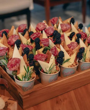 Bite-sized charcuterie cups on a wooden tray with cheese wedges, rolled salami, crispy crackers, fresh blackberries, rosemary sprigs and mixed nuts — elegant party appetizers.