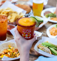 Hand holding a frozen mango cocktail with a chili-salt rim and lime wedge, surrounded by tacos, chips, salsa and a beer on a wooden picnic table — vibrant Mexican street-food spread.