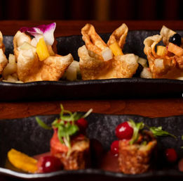 Close-up of gourmet Asian-fusion appetizers: crispy fried wonton cups filled with diced mango and apple and topped with an edible orchid on a black serving tray, with bite-sized fried rolls garnished with microgreens and bright red berries in the foreground.