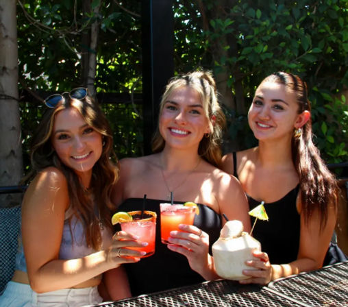 Three friends smiling on a sunny outdoor patio, holding pink citrus cocktails and a coconut drink with a tiny umbrella, with leafy greenery in the background.