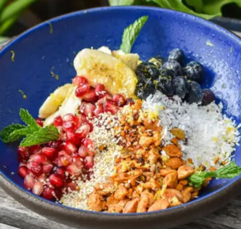 Colorful fruit-and-nut breakfast bowl in a blue dish with banana slices, blueberries, pomegranate arils, shredded coconut, chopped nuts, seeds and mint on a wooden table