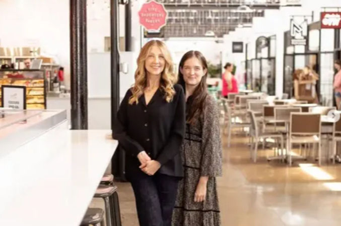 Two smiling women pose by a long white counter inside a bright indoor food hall, with tables, chairs and vendor stalls visible in the background.