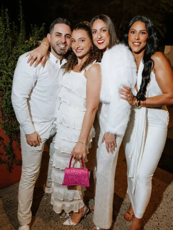 Group of four adults in white outfits smiling at an outdoor evening party — women in white dresses (one with a fluffy jacket) and a man in a white shirt; one woman holds a small pink glitter handbag.