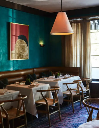 Cozy bistro dining corner with emerald teal walls, leather banquette, wooden wishbone chairs, white-clothed tables set for service, oversized cone pendant light, sheer window curtains and patterned rug.