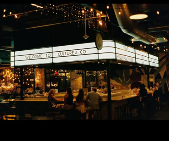 Cozy urban food-hall bar at night with a lighted marquee welcome sign above a curved central counter, warm string lights and patrons seated at the bar