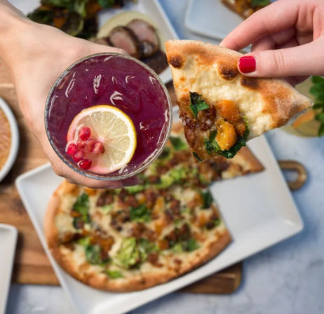 Hands holding a pomegranate‑colored cocktail with a lemon slice and pomegranate seeds alongside a hand grabbing a slice of roasted‑vegetable flatbread pizza on a white plate at a casual restaurant table