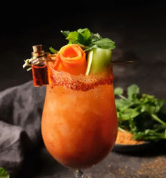 Spicy Bloody Mary cocktail in a stemmed glass with a chili-rimmed edge, carved carrot rose, cucumber ribbon and cilantro with a tiny hot-sauce vial garnish on a dark tabletop