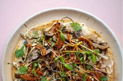 Overhead shot of a gourmet mushroom flatbread on a ceramic plate—mixed sautéed mushrooms, crispy shallots, microgreens and cilantro on a creamy spread, cafe-style on a pink tabletop.