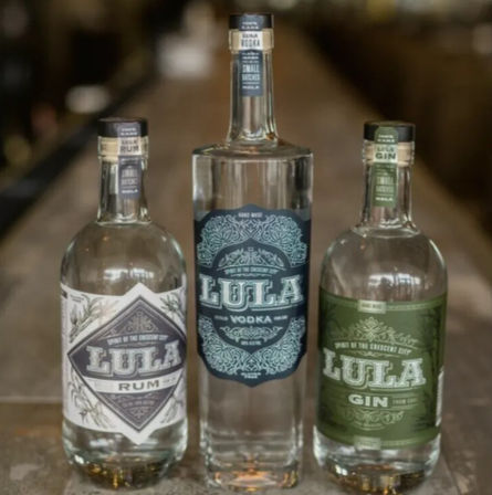Three clear craft spirit bottles—Lula vodka (center), Lula rum (left) and Lula gin (right)—standing on a rustic wooden bar counter with ornate decorative labels.