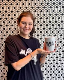 Smiling person holding a white 'Milk & Honey' coffee mug against a black-and-white hexagon tile wall in a cozy cafe-style scene.