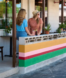 Two people chatting over drinks at a colorful striped cinderblock patio wall of an outdoor cafe with potted plants on a sunny sidewalk