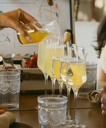 Hand pouring bright orange juice into a row of champagne flutes to make mimosas on a brunch table with patterned water glasses.
