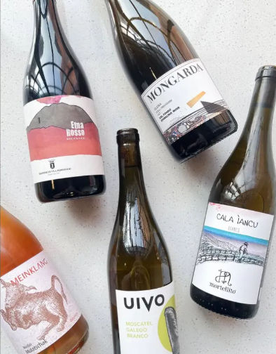 Five assorted wine bottles—red, white and rosé—arranged on a light stone countertop with colorful labels, evoking an artisanal wine tasting selection.