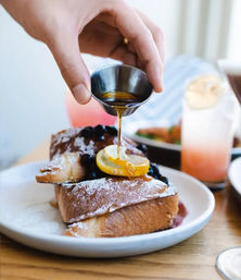 Hand pouring maple syrup from a metal cup onto powdered French toast stacked with blueberries and a lemon slice on a white plate at a brunch table.