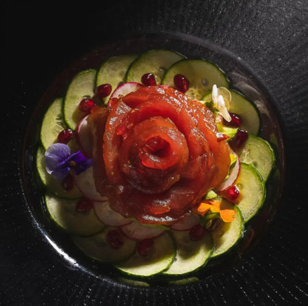 Sashimi rose of thinly sliced raw fish on cucumber rounds, garnished with pomegranate arils and edible flowers on a black plate — elegant sushi-style fine-dining presentation.