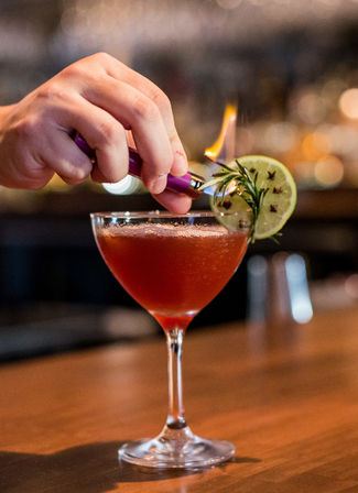 Hand torching a rosemary garnish on a ruby cocktail in a coupe glass, garnished with a clove-studded lime slice on a wooden bar top with blurred bokeh lights