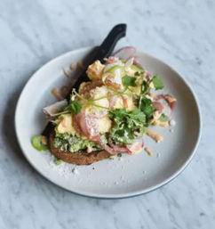 Cafe-style brunch avocado toast: smashed avocado and two poached eggs with pickled radishes, cilantro, grated cheese and creamy sauce on toasted sourdough, served with a black-handled knife on a white plate over a marble table.