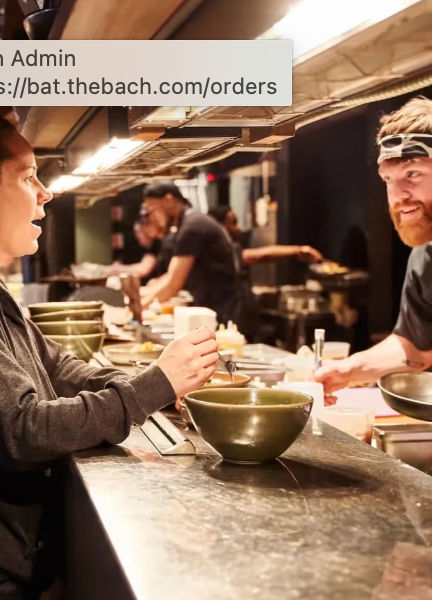 Busy open-kitchen restaurant pass with a chef handing a large green bowl to a customer across a marble counter, other cooks plating dishes in the background.