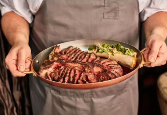 Hands holding a copper pan of sliced grilled steak with charred sear marks and roasted bone marrow topped with herbs, rustic steakhouse presentation.