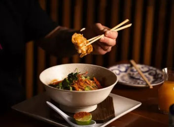 Chopsticks lifting a dumpling above an Asian noodle bowl with greens and shredded carrots, lime half and spoon on a square plate, patterned side plate and juice glass in a cozy wood-paneled restaurant setting.