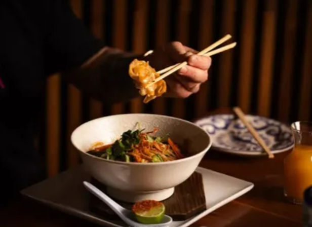 Chopsticks lifting a dumpling above an Asian noodle bowl with greens and shredded carrots, lime half and spoon on a square plate, patterned side plate and juice glass in a cozy wood-paneled restaurant setting.