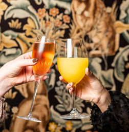 Hands clinking a pink sparkling cocktail and a bright orange mimosa in stemmed glasses against decorative floral wallpaper — brunch toast