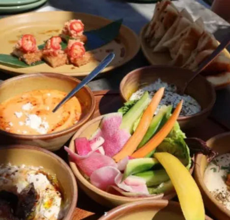 Sunlit patio table with a colorful mezze spread: bowls of creamy dips, pita wedges and crostini, plus fresh crudités—cucumber, carrot sticks, radish slices and lettuce.