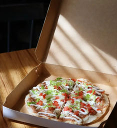 Sunlit thin-crust takeout pizza in an open cardboard box on a wooden table, topped with diced tomatoes, shredded lettuce, cheese and drizzled white sauce.