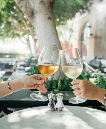 Two hands clinking a rosé and a white wine glass over a sunlit outdoor café table with salt-and-pepper shakers and leafy patio background.