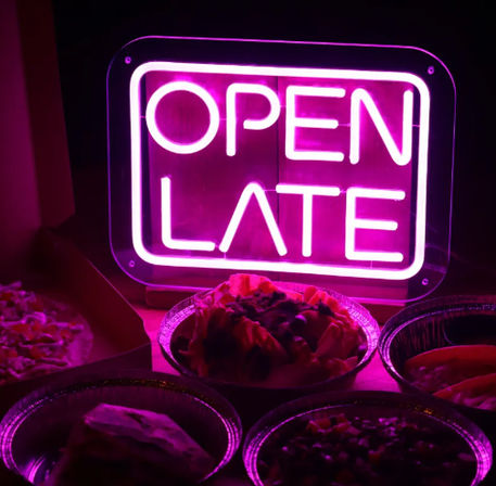 Glowing pink neon "OPEN LATE" sign illuminating assorted late-night takeout bowls and a pizza slice in a dim restaurant setting