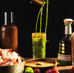Bright green tequila cocktail being poured into a tall textured glass with a spicy-salt rim, surrounded by shrimp, spices, chiles and a tequila bottle on a dark background.