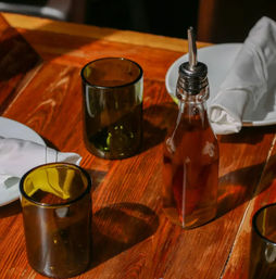 Rustic wooden dining table with a glass olive oil cruet with pourer, amber drinking tumblers, white rolled napkins and plates