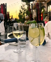 Al fresco patio dining at sunset — white wine glass and lime-mint spritzer with straw on a marble table, string lights and blurred terrace crowd in the background.