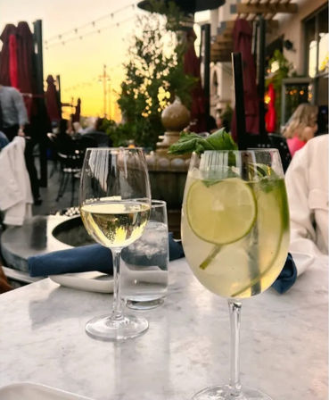 Al fresco patio dining at sunset — white wine glass and lime-mint spritzer with straw on a marble table, string lights and blurred terrace crowd in the background.