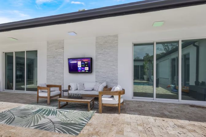 Sunny modern covered patio with wooden lounge set and white cushions, tropical outdoor rug, wall-mounted TV, and sliding glass doors reflecting a pool — stylish backyard living space.
