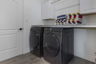 Bright residential laundry room with matching black front-load washer and dryer under a granite countertop, white wall cabinets above, neatly folded colorful striped towels and gray wood-look flooring.