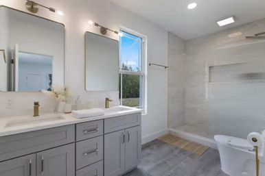 Bright, airy modern bathroom with gray double-sink vanity, gold faucets and two mirrors, window with outdoor view, glass walk-in shower and white toilet on gray tile floor.