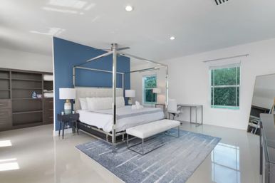 Bright modern master bedroom with chrome four‑poster canopy bed, blue accent wall, white bedding, bench on a gray-blue rug, desk by large windows overlooking green yard and open walk-in closet