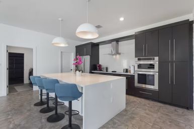Bright contemporary kitchen with large white island, four blue swivel bar stools, dark floor-to-ceiling cabinetry, stainless-steel ovens and range hood, pendant lights and pink orchid centerpiece.