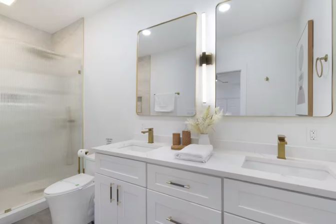 Bright modern white bathroom with double vanity and quartz countertop, gold faucets and framed mirrors with vertical lights, frosted glass walk‑in shower, toilet, folded towels and decorative vase.
