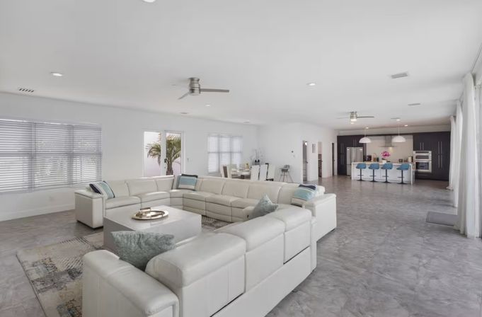 Bright, spacious open-plan living area with oversized white leather sectional, central coffee table, large windows and sliding doors, gray polished floors, dining area and modern kitchen island with blue bar stools.