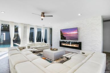 Sunlit modern living room with large white sectional sofas around a square coffee table, wall-mounted TV displaying a sunset above a linear fireplace, floor-to-ceiling windows with sheer curtains and a view of a backyard pool, stacked-stone accent wall and ceiling fan.
