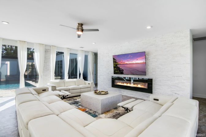 Sunlit modern living room with large white sectional sofas around a square coffee table, wall-mounted TV displaying a sunset above a linear fireplace, floor-to-ceiling windows with sheer curtains and a view of a backyard pool, stacked-stone accent wall and ceiling fan.