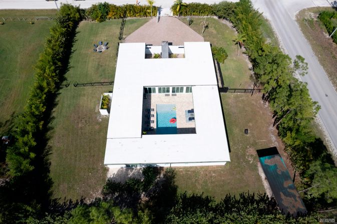 Drone aerial view of a modern white-roofed U-shaped house with a central courtyard pool, lounge chairs and red float, set on a grassy lot bordered by trees and a nearby road.