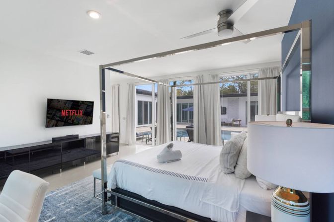 Bright modern master bedroom with chrome four‑poster bed, white linens and folded towel, wall‑mounted TV showing Netflix, sliding glass doors revealing a private pool patio, plush blue rug and bedside lamp