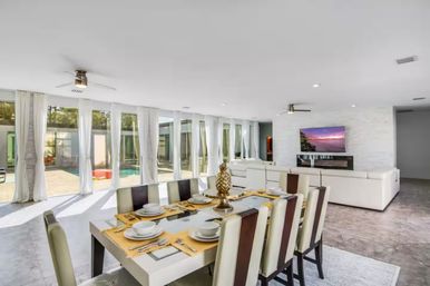 Bright modern open-plan dining and living area with a table set for eight, white leather sofas, ceiling fans, floor-to-ceiling glass doors and a sunlit pool patio visible outside.