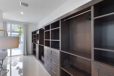 Sleek modern walk-in closet with dark wood built-in shelving and drawers, chrome hanging rods, glossy white tile floor, and a sliding glass door with sheer curtains opening to a sunlit backyard pool
